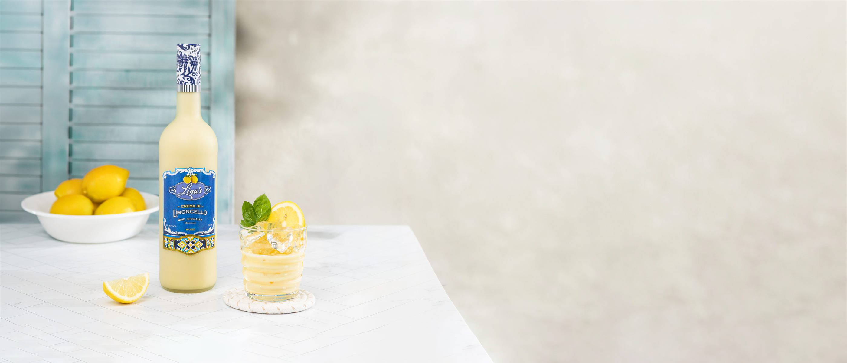 A bottle of Lina's Crema di Limoncello beside a glass over ice and a bowl of lemons